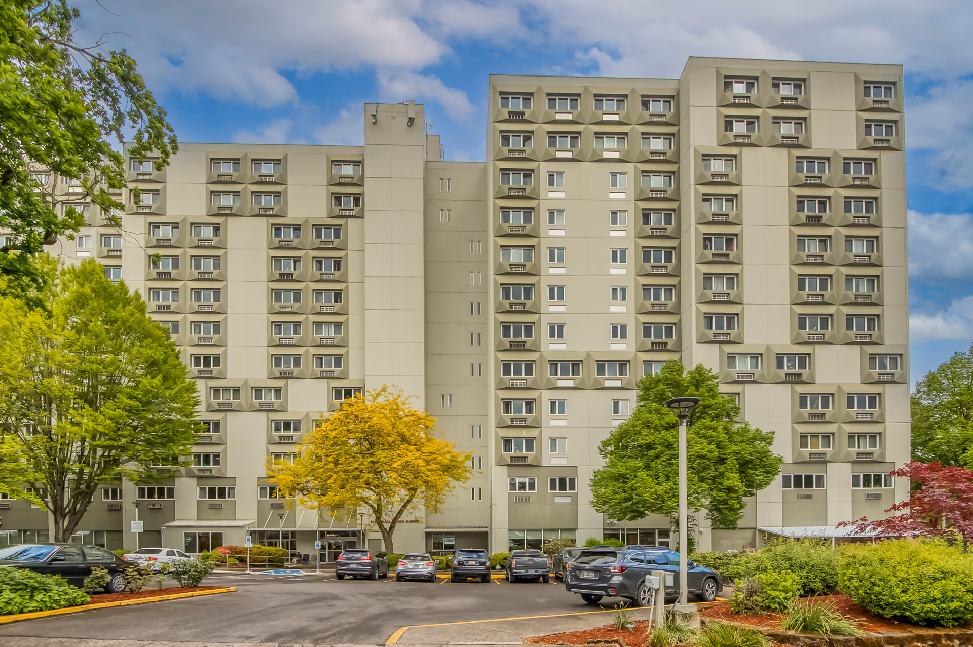 Exterior view of a multi-story senior living facility building with numerous windows, surrounded by trees and parked cars in the foreground under a partly cloudy sky.