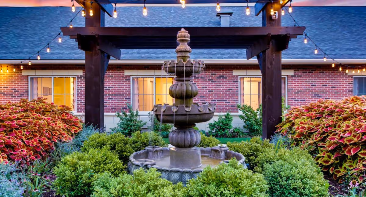 Outdoor garden area at Waverly Inn Memory Care Community featuring a multi-tiered stone fountain surrounded by green bushes and colorful plants, with a brick building and string lights in the background.