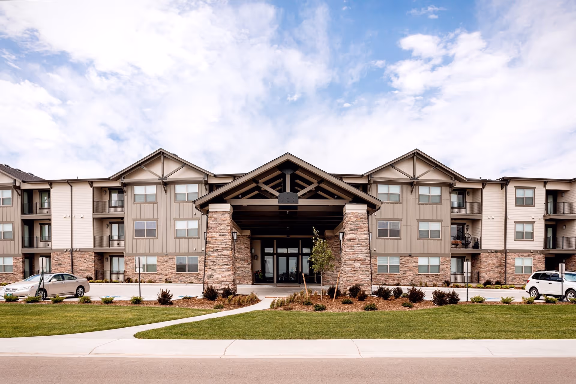 Front exterior view of a three-story senior living facility building named Eagle's Peak with stone and beige siding, a covered entrance supported by stone pillars, landscaped greenery, and parked cars on either side.