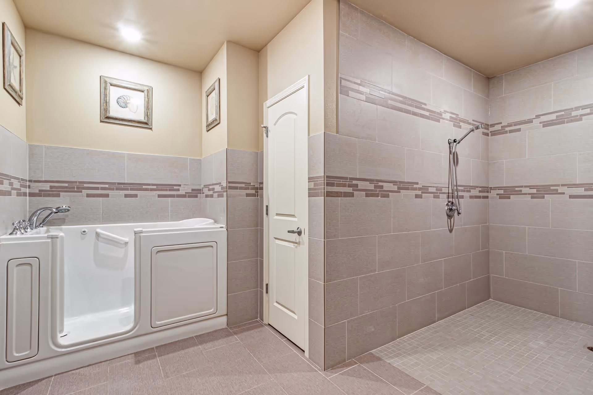 A spacious bathroom featuring a walk-in bathtub with a door on the left side and a large open shower area with a handheld showerhead on the right. The walls and floor are tiled in neutral tones with decorative horizontal tile accents. There is a white door in the center and framed artwork on the walls above the bathtub. The room is well-lit with recessed ceiling lights.