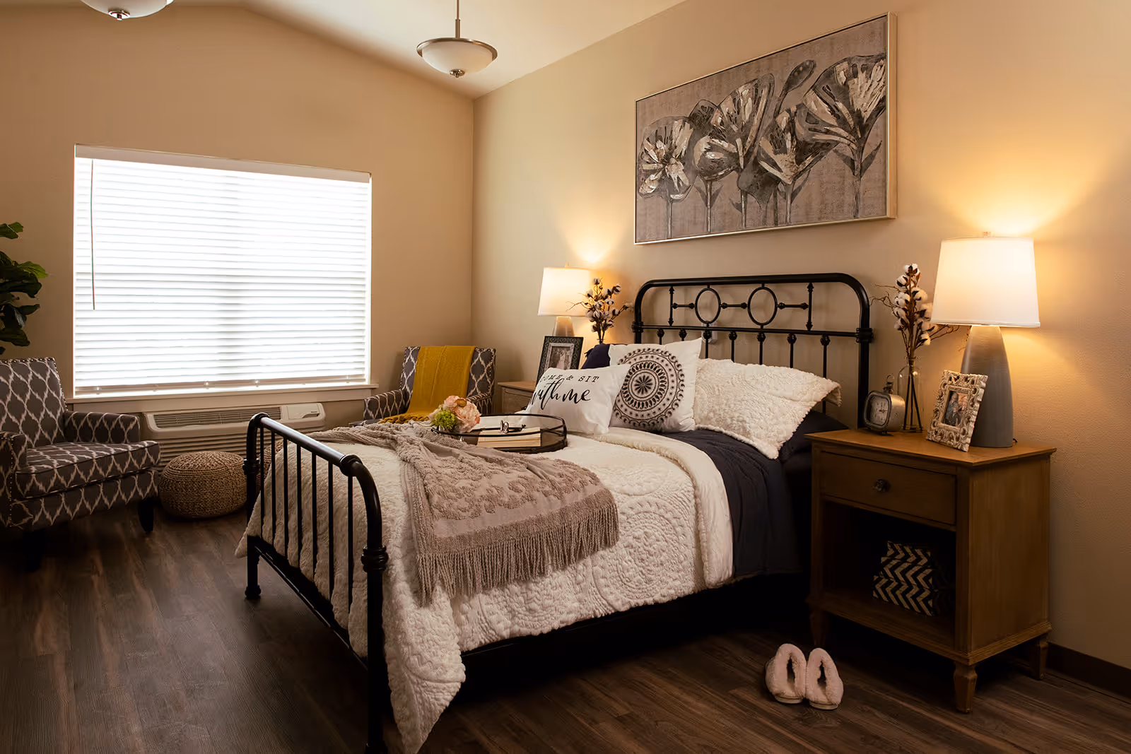 A cozy bedroom with a black metal bed frame, white and beige bedding, and decorative pillows. There are two wooden nightstands with lamps and framed photos on each side of the bed. Two patterned armchairs and a small round ottoman are placed near a window with closed blinds. A large floral artwork hangs above the bed, and a pair of slippers is on the wooden floor near the nightstand.