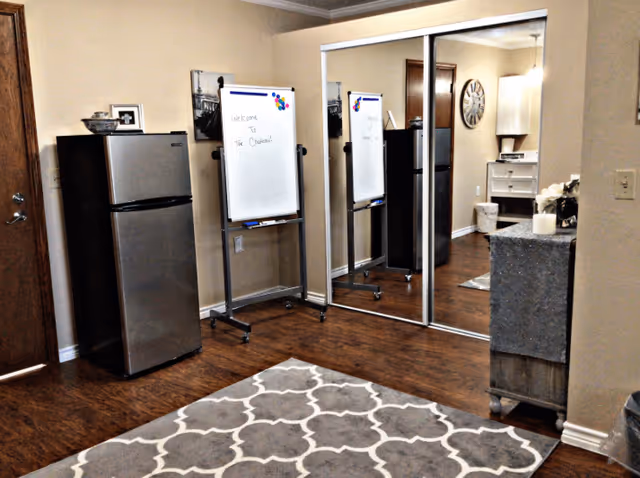 Interior room with wooden floor, a small stainless steel refrigerator, a whiteboard on a stand with the message 'Welcome to The Chateau!', mirrored sliding closet doors, a patterned area rug, and a small cabinet with decorative items on top.