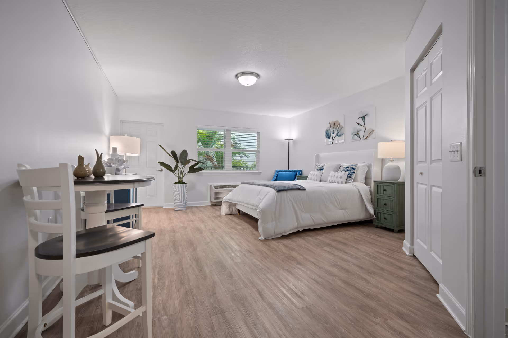 A bright and spacious bedroom with a large bed dressed in white linens and decorative pillows. The room features light wood flooring, a small round table with two chairs, a green nightstand with a lamp, a tall floor lamp, and a potted plant near a window with blinds. The walls are white with two floral artworks above the bed.