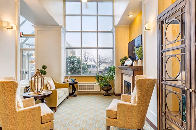 A bright and cozy living room area with large floor-to-ceiling windows letting in natural light. The room features two upholstered armchairs, a sofa, a wooden side table with decorative items, a fireplace with a TV mounted above it, and several potted plants. The walls are painted yellow and white, creating a warm and inviting atmosphere.