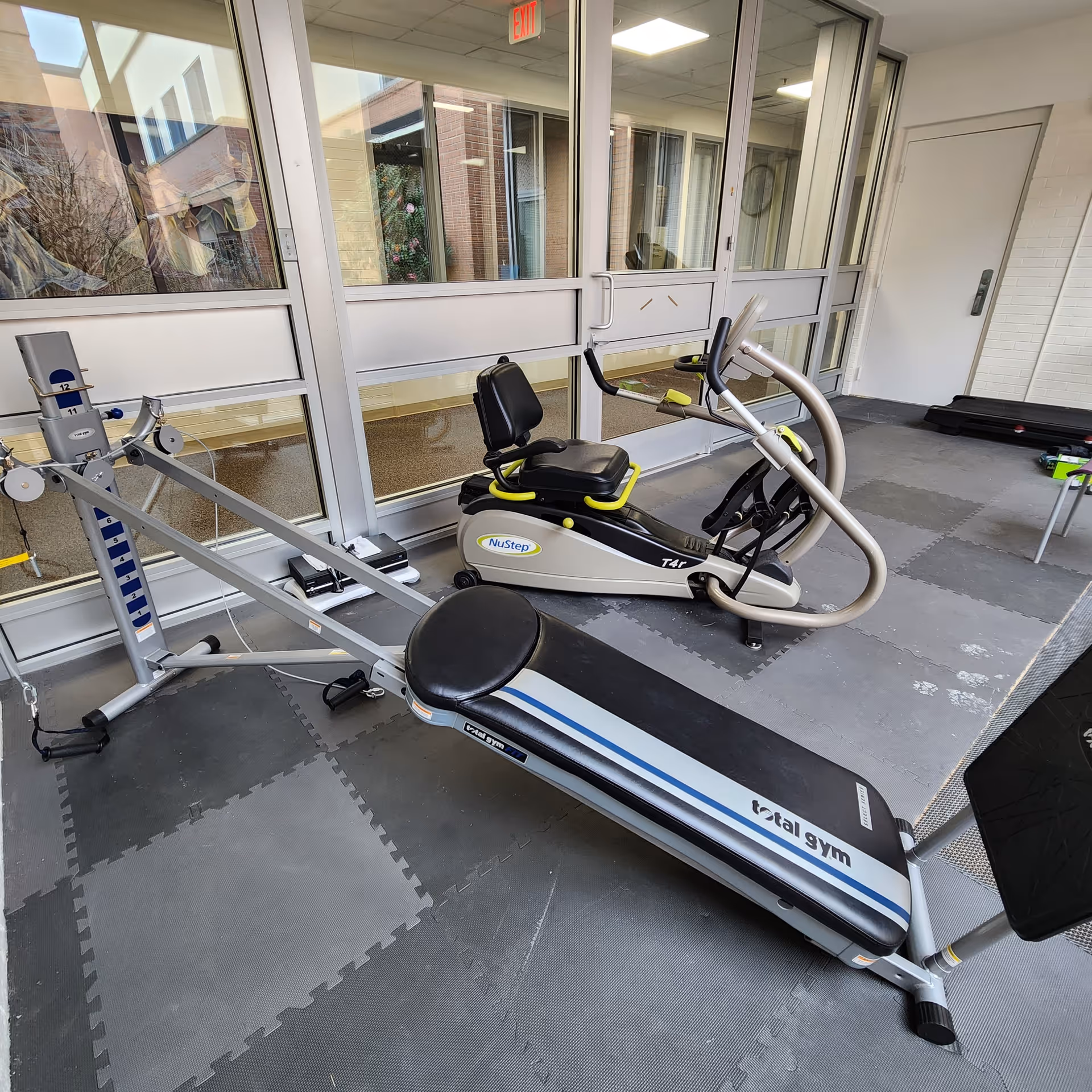 Indoor exercise area with fitness equipment including a NuStep recumbent cross trainer and a Total Gym incline workout bench on gray interlocking foam floor mats. The room has large windows and a white door in the background.