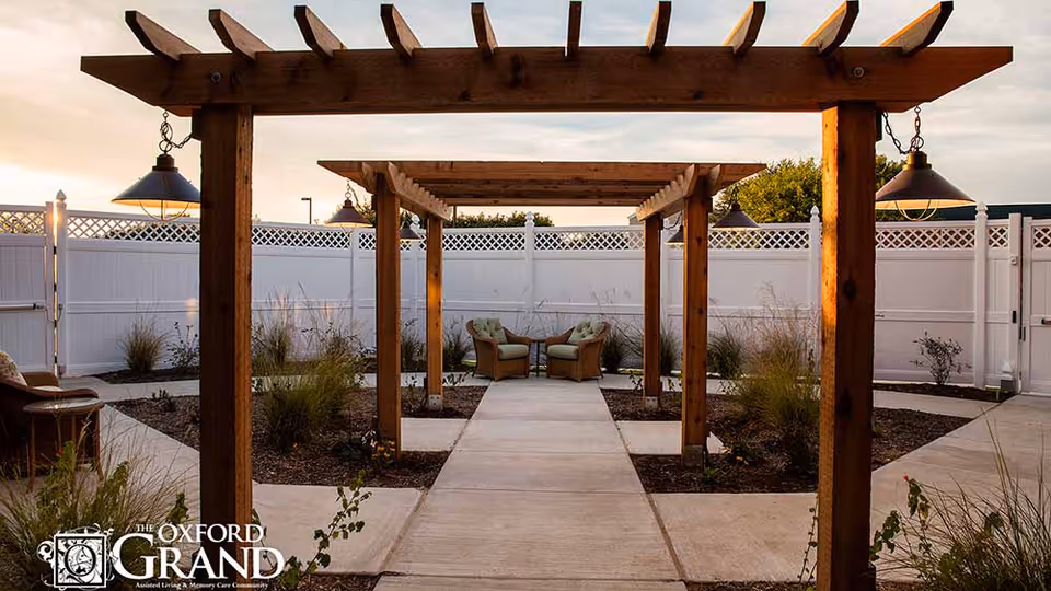 Outdoor courtyard with wooden pergolas, a paved walkway, and two cushioned chairs in front of a white fence.