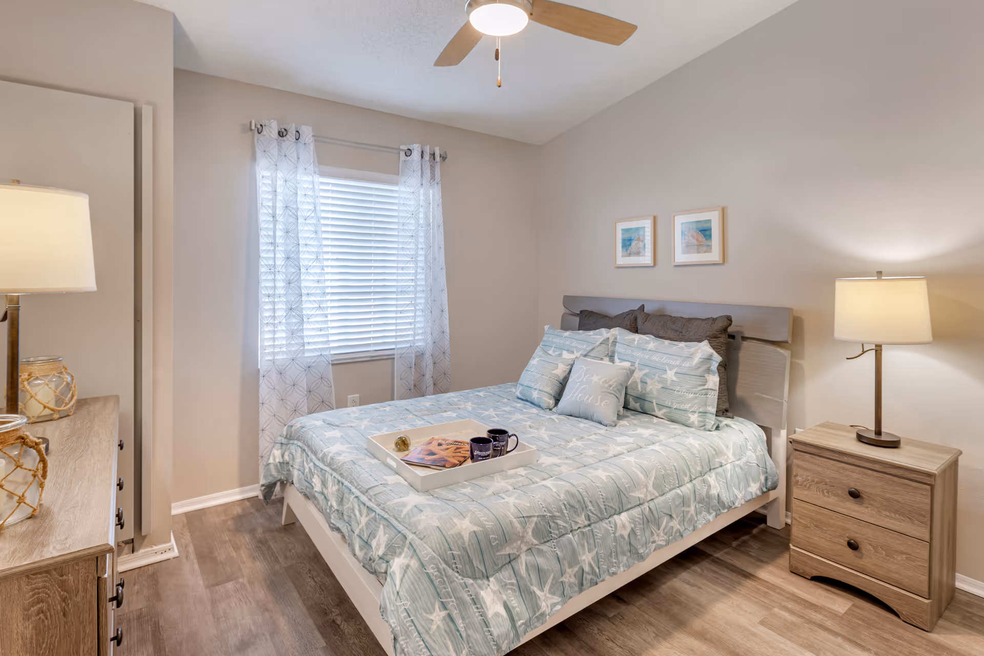 A cozy bedroom with a bed dressed in light blue and white starfish patterned bedding, two framed pictures on the wall above the headboard, a wooden nightstand with a lamp on the right side, a wooden dresser with a lamp on the left, and a window with white blinds and sheer patterned curtains.