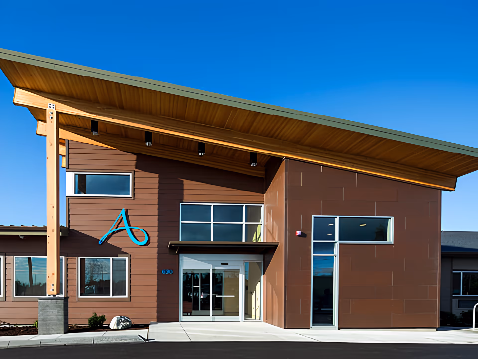 Exterior front view of Avamere Transitional Care of Puget Sound building with modern architecture, large windows, and a wooden overhanging roof under a clear blue sky.