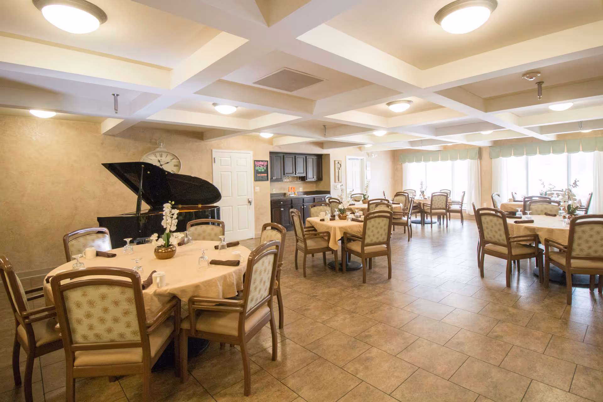 A spacious dining room with multiple round tables covered with beige tablecloths, each surrounded by wooden chairs with cushioned seats and backs. The room features a tiled floor, a grand piano against one wall, a large clock, and a small kitchenette area with cabinets and a countertop. Large windows with light green valances allow natural light to fill the room.
