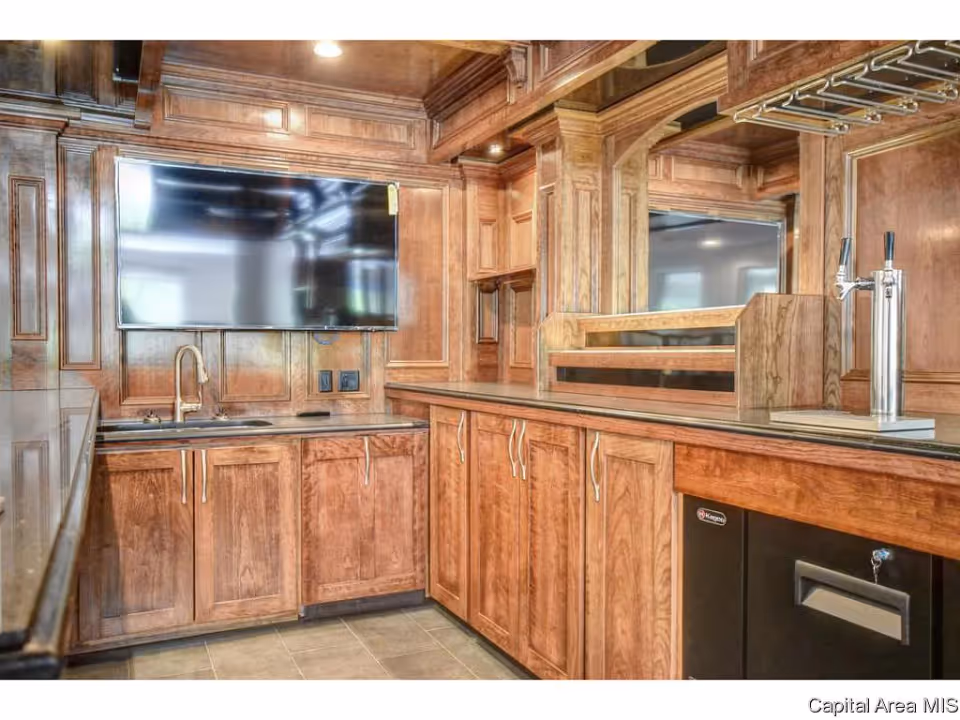 Interior view of a wooden kitchenette or bar area featuring multiple cabinets with silver handles, a sink with a gold faucet, a mounted flat-screen TV, a beer tap, and a mirrored backsplash.