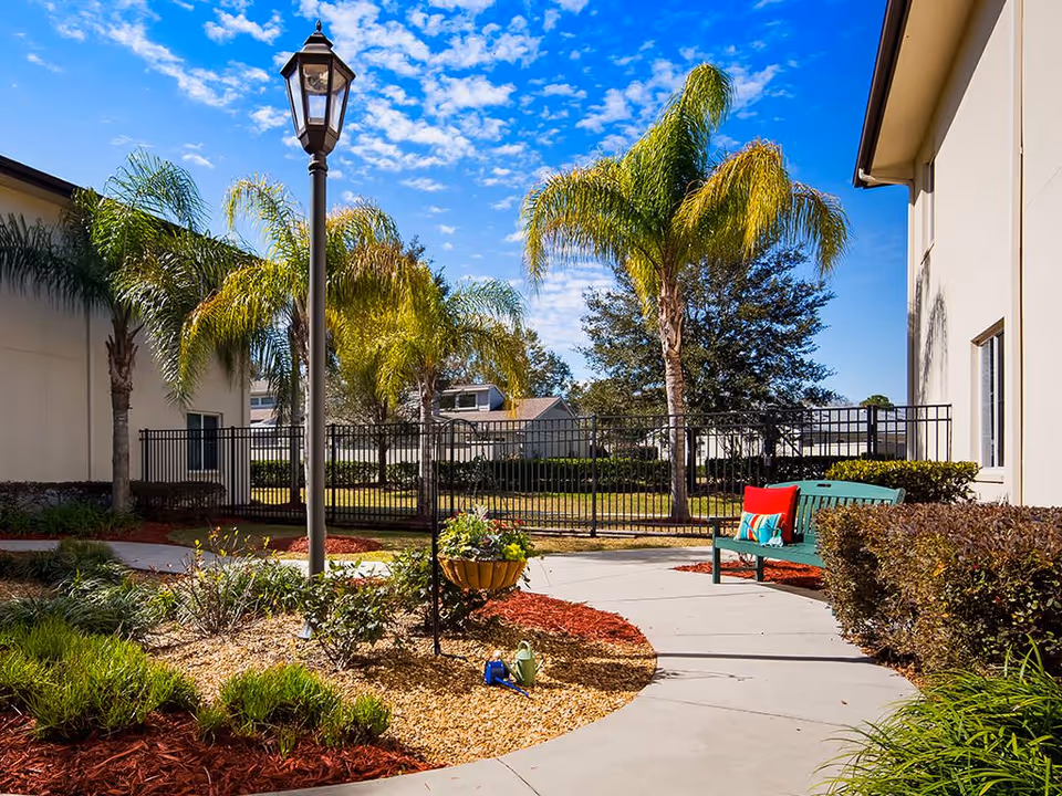 Outdoor courtyard area with a curved concrete pathway, green bench with colorful cushions, palm trees, a black metal fence, a lamp post, and landscaped garden beds with mulch and plants under a blue sky with scattered clouds.