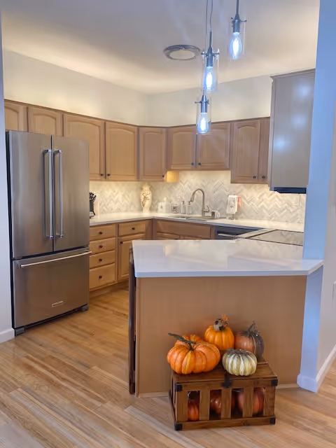 Modern kitchen with light wood cabinets, stainless steel refrigerator, white countertops, and a herringbone tile backsplash. Three pendant lights hang from the ceiling. A wooden crate with decorative pumpkins is placed on the floor near the kitchen island.