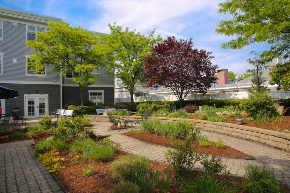 A landscaped courtyard with paved walkways, benches, tables, and trees beside a multi-story building.