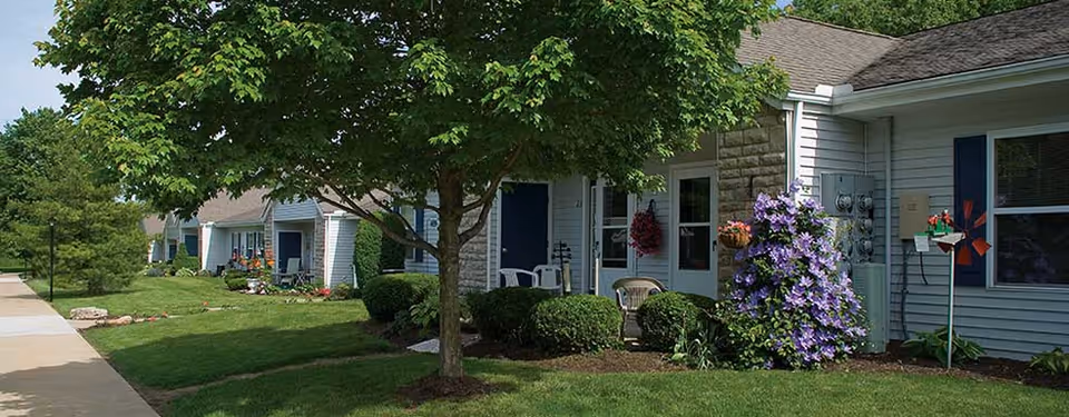 A row of single-story residential units with light blue siding and dark blue doors, each with a small porch area. The units are surrounded by well-maintained green lawns, bushes, and trees, with a concrete sidewalk running alongside the buildings. There are flowers and decorative plants near the entrances, and the sky is clear and blue.