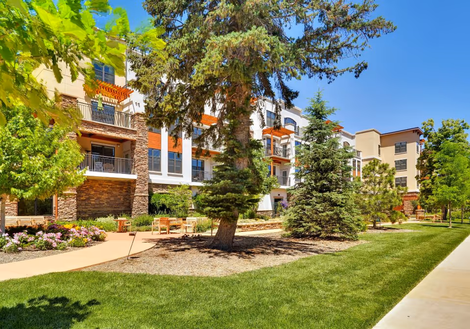 Outdoor view of The Pearl at Boulder Creek facility showing a landscaped garden with green grass, trees, and flower beds. The multi-story building with balconies and stone accents is visible in the background under a clear blue sky.