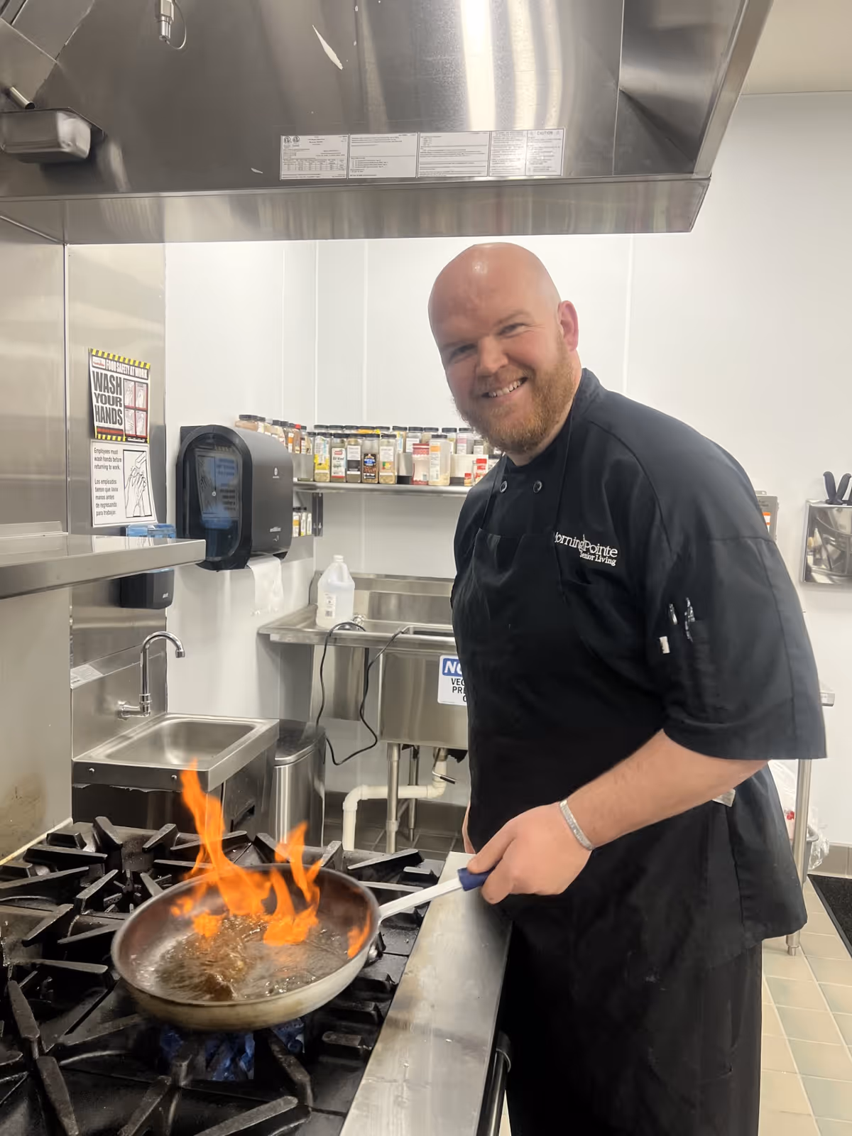 A smiling chef wearing a black Morning Pointe uniform is cooking with a flaming pan on a gas stove in a commercial kitchen. Shelves with spices and kitchen supplies are visible in the background.