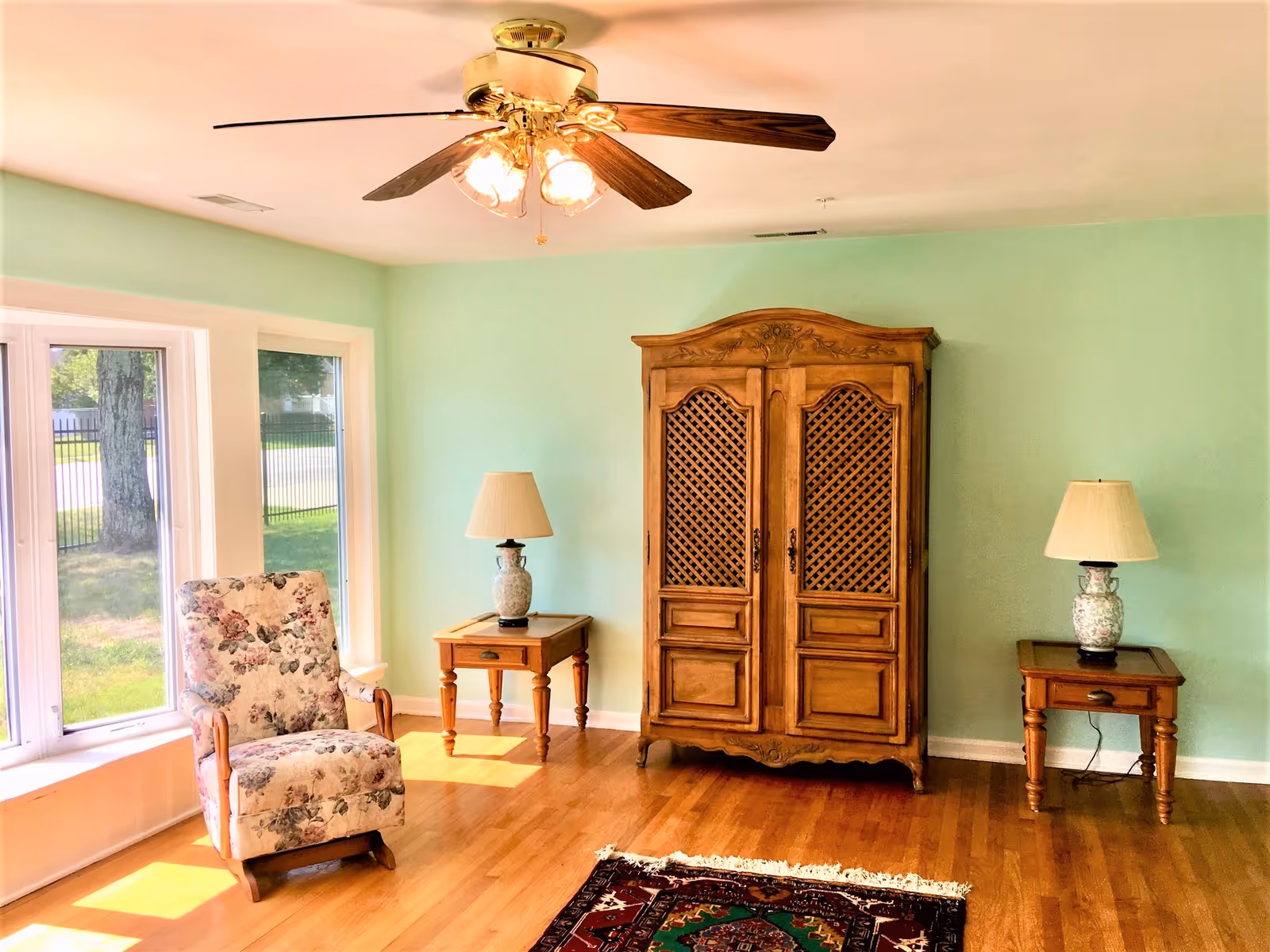 Sunlit living room with a floral armchair by large windows, a wooden armoire, two end tables with lamps, a ceiling fan, and a small rug on hardwood floors.