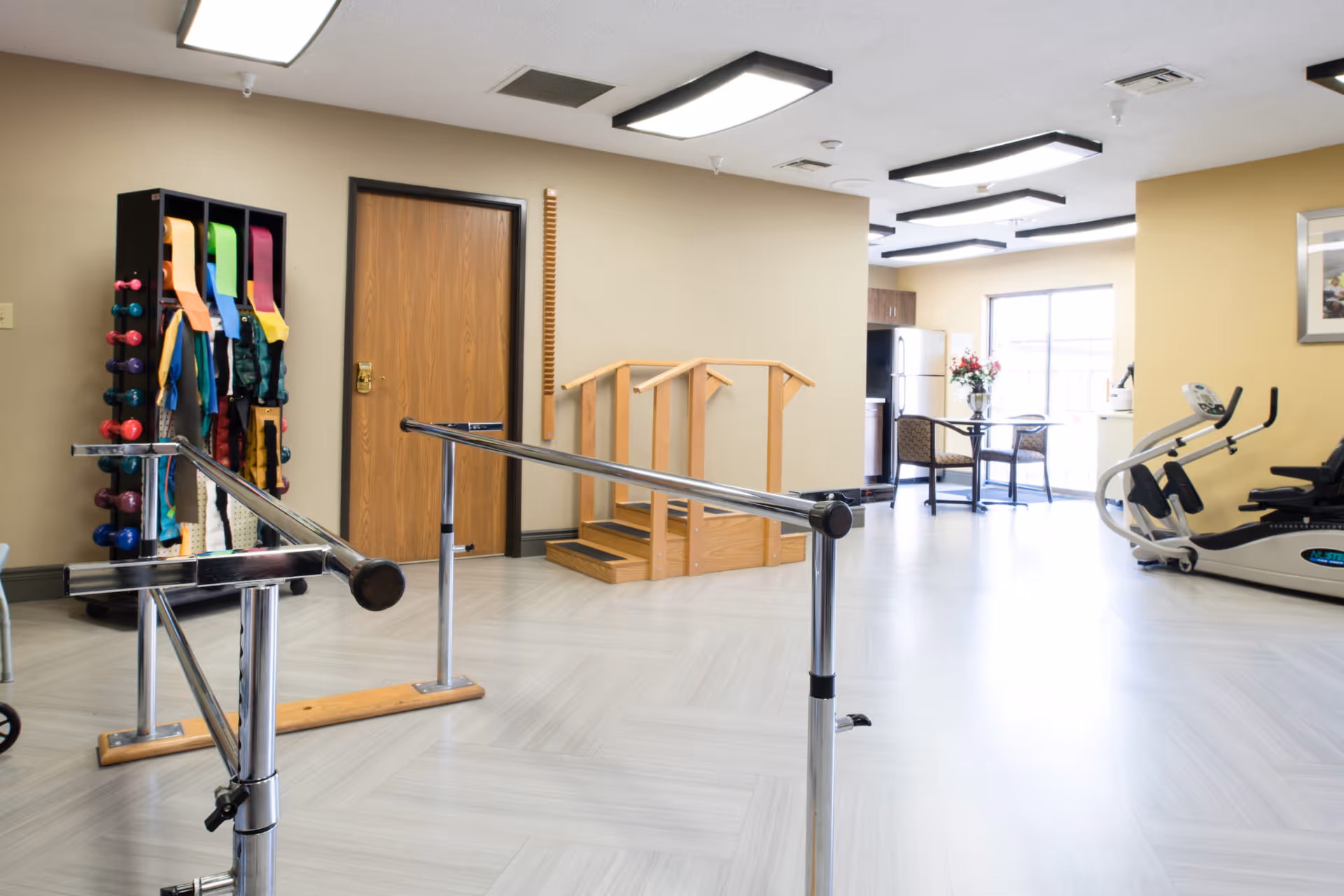 A senior living facility exercise room with parallel bars for walking support, a rack holding colorful exercise bands and dumbbells, a wooden step platform with handrails, an exercise bike, and a small table with two chairs near a window letting in natural light.