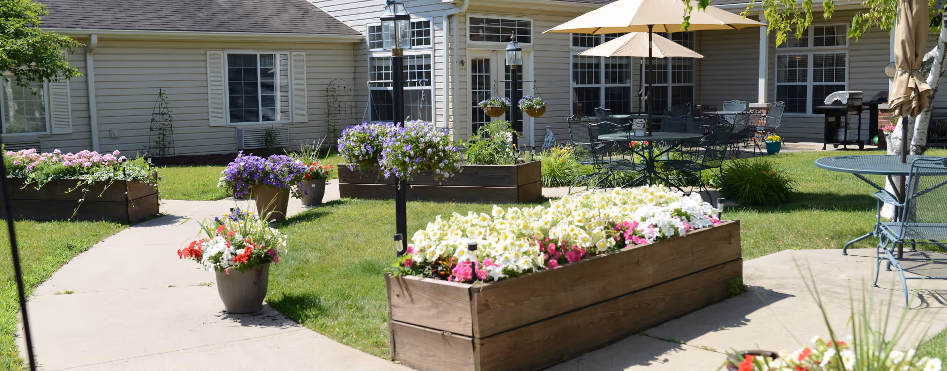 Outdoor courtyard area at Bickford of Sioux City featuring raised wooden flower beds filled with colorful flowers, green grass, paved walkways, patio tables with umbrellas, and a building with windows and siding in the background.