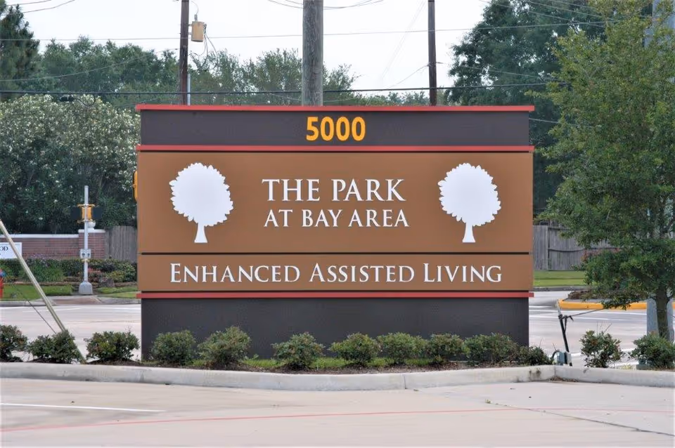 Roadside sign for The Park At Bay Area enhanced assisted living with address number 5000 and tree logos in front of landscaping and a street.