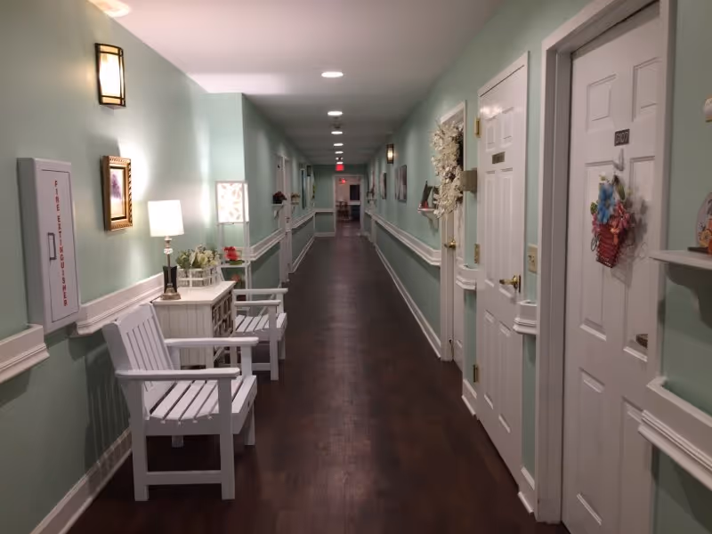 A long, well-lit hallway in an assisted living facility with light green walls, dark wood flooring, and white trim. There are white doors on the right side, some decorated with wreaths and flowers. On the left side, there are white chairs, a small white table with a lamp and flowers, wall-mounted lights, and framed pictures. The hallway extends into the distance with ceiling lights and an exit sign visible at the far end.