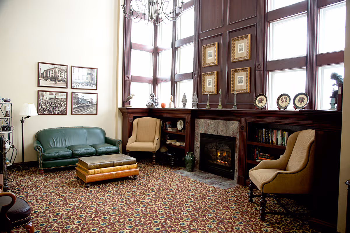 A cozy living room with a green leather sofa, two beige armchairs, a fireplace with a wooden mantel, and large windows letting in natural light. The room features a patterned carpet, framed pictures on the walls, and decorative items on the mantel and shelves.
