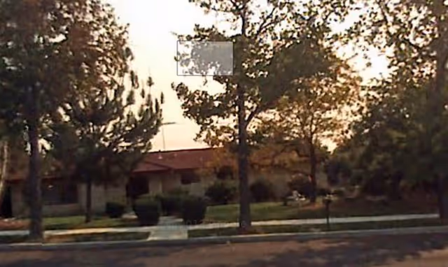 View of a single-story building partially obscured by trees and bushes, with a sidewalk and street in the foreground during sunset or early evening.