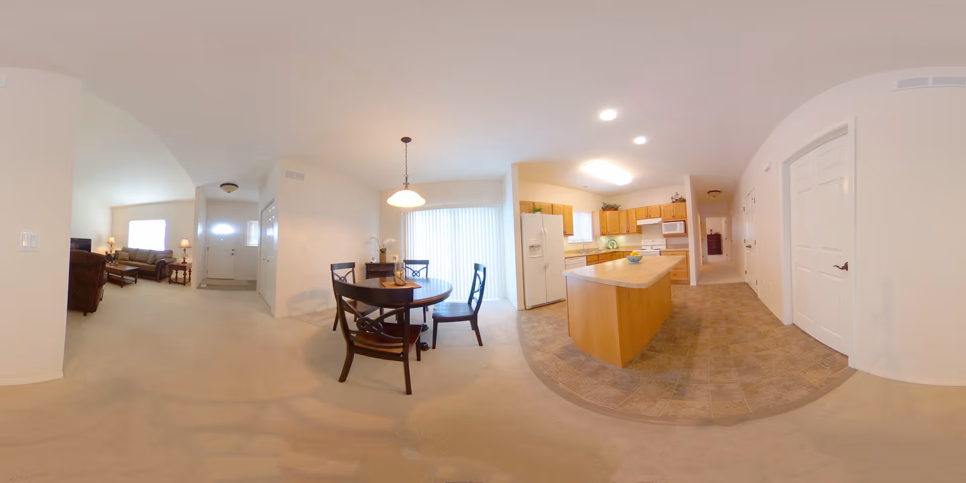 Wide-angle view of an open floor plan in a senior living facility showing a kitchen with wooden cabinets, a central island, and appliances including a refrigerator and stove. Adjacent to the kitchen is a dining area with a round wooden table and four chairs. Beyond the dining area is a living room with a sofa, armchair, and lamps. The space is well-lit with natural light coming through a large window with vertical blinds.