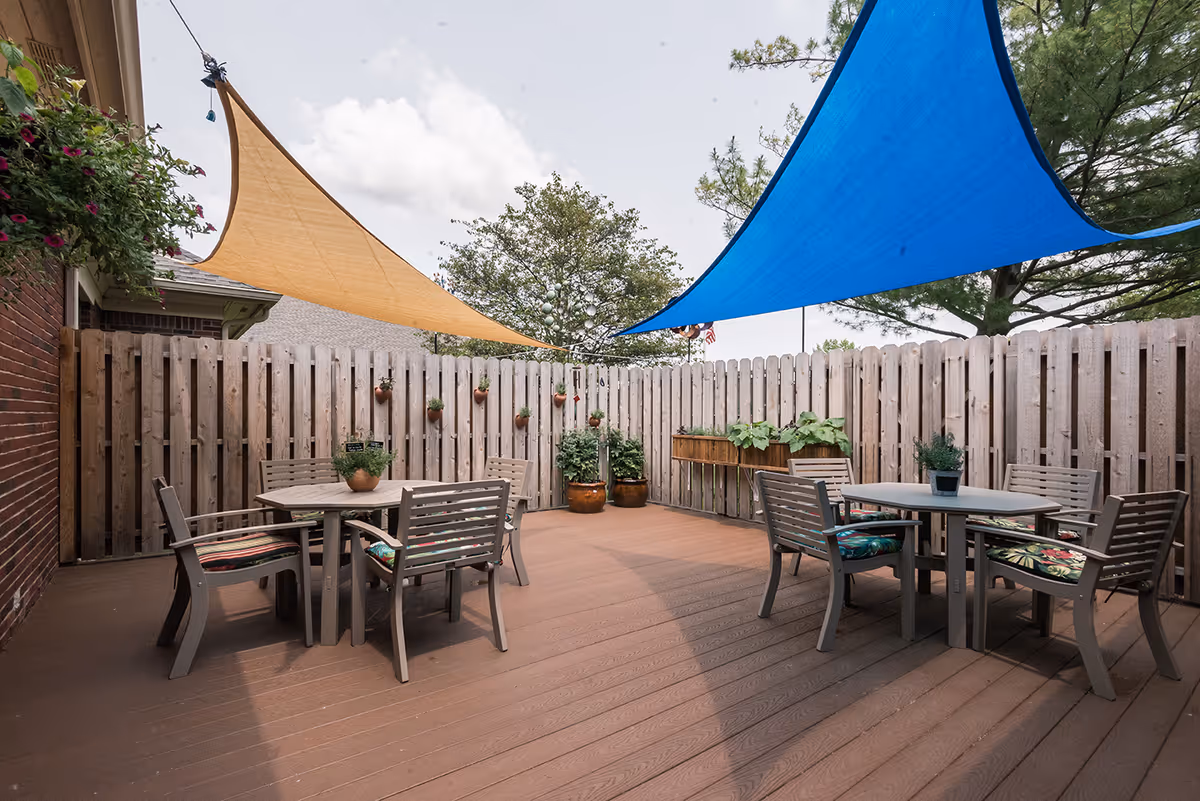 Outdoor fenced patio with two dining tables, chairs, potted plants and colorful shade sails overhead.