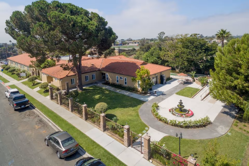 Aerial view of Arroyo Vista Nursing Center showing a single-story building with a red tile roof surrounded by green lawns, trees, and a circular driveway with a central fountain. Several cars are parked along the street adjacent to the facility.
