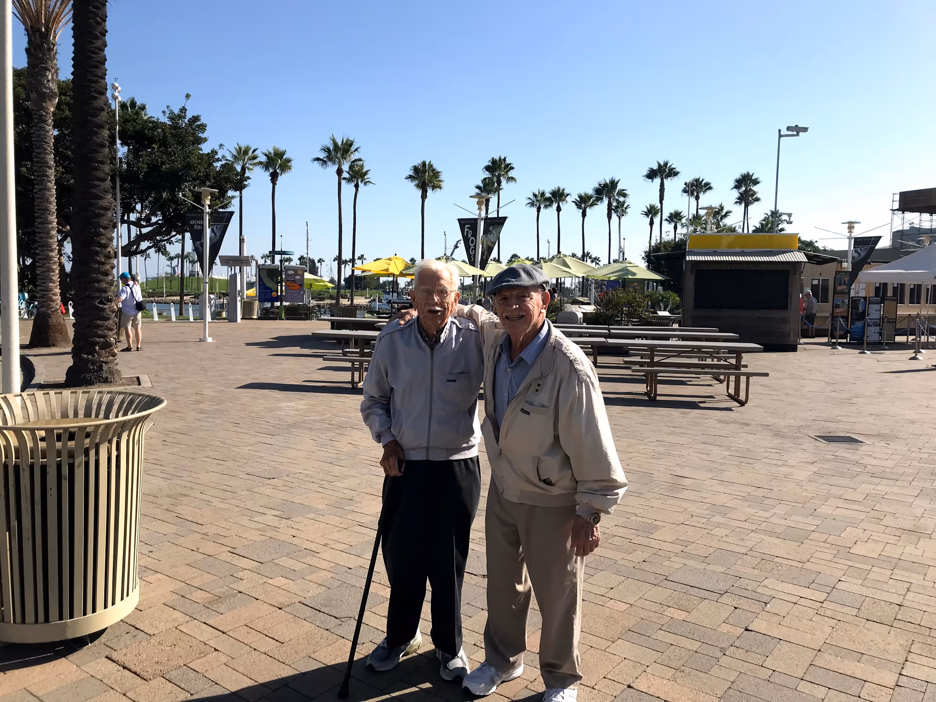 Two elderly men standing outdoors on a paved area with palm trees and picnic tables in the background. One man is using a cane and the other has his arm around him. The sky is clear and blue.