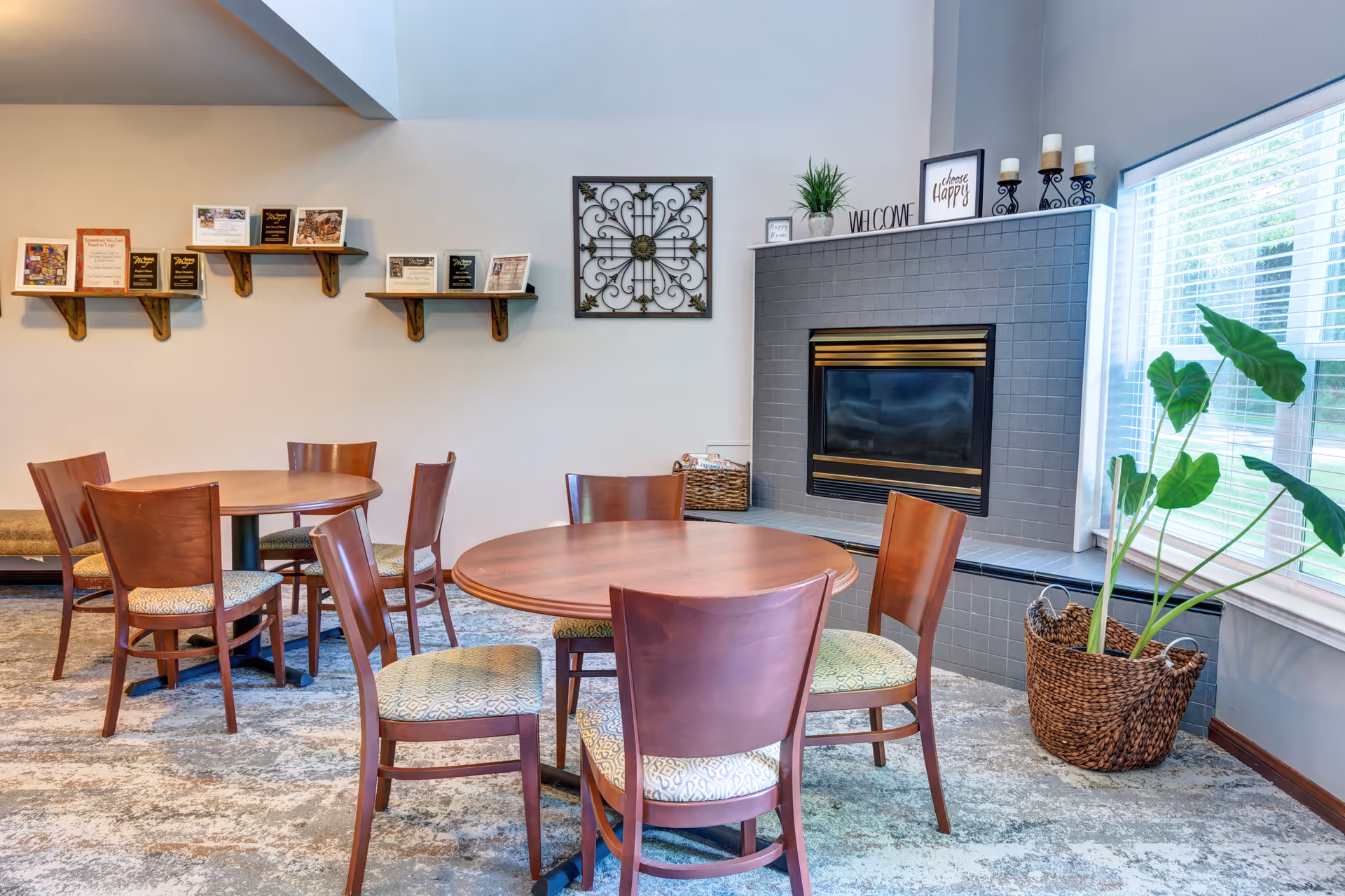 A cozy common area with round wooden tables and chairs arranged on a carpeted floor. There is a gray tiled fireplace with decorative items on the mantel, including candles, a plant, and framed signs. A large window with white blinds lets in natural light, and a woven basket with a green plant sits beside the fireplace. Shelves on the wall display plaques and awards.