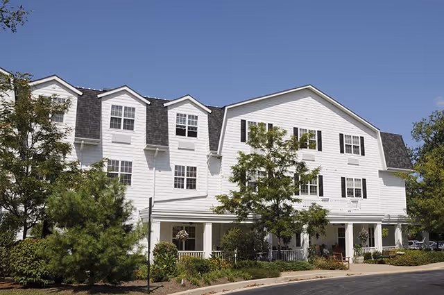 Exterior view of a large white senior living facility building with multiple windows and a covered porch area. The building is surrounded by trees and landscaped greenery under a clear blue sky.