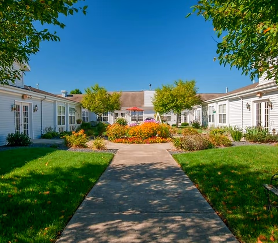 A paved walkway leads through a landscaped courtyard with colorful flowerbeds and single-story white buildings on either side under a clear blue sky.