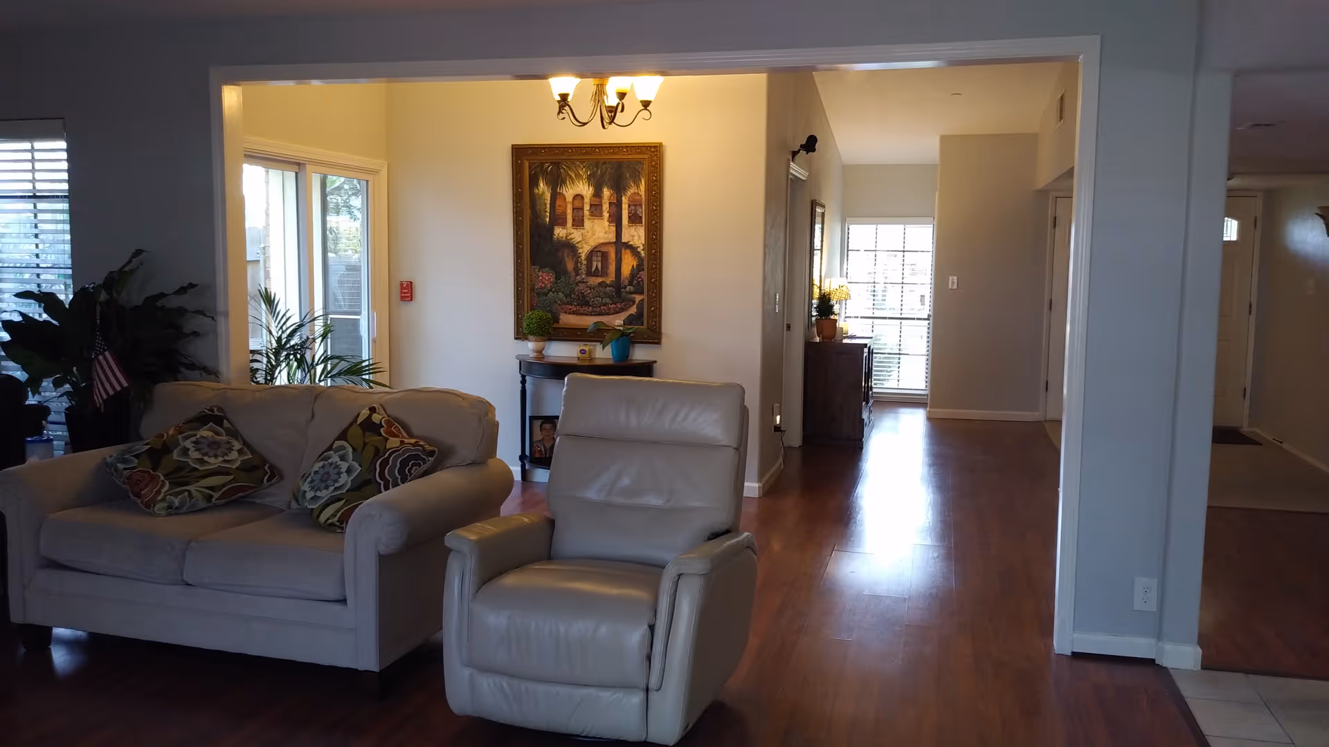 Open living room with a beige sofa, a leather recliner, decorative pillows, plants, artwork, and a hallway leading to other rooms.