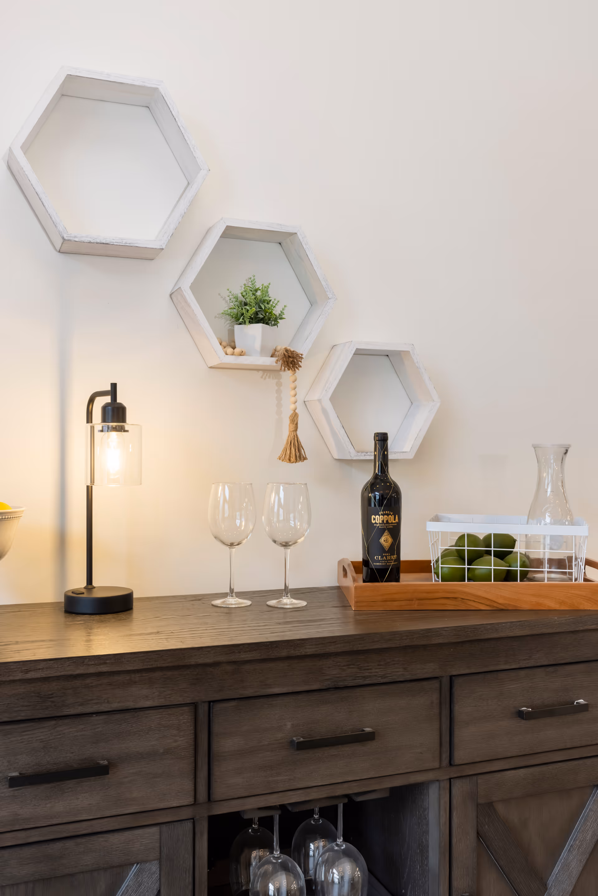 A wooden sideboard with a modern black lamp, two empty wine glasses, a bottle of Coppola wine, and a wooden tray holding a white wire basket with green apples and a glass pitcher. Above the sideboard are three white hexagonal wall shelves, one holding a small potted plant and a decorative tassel.