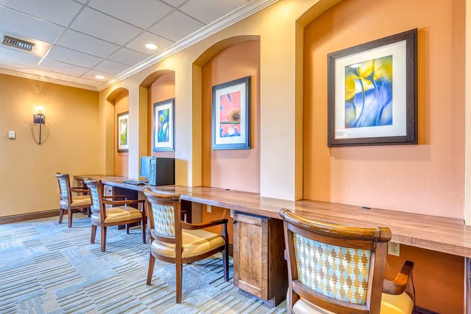A well-lit interior room with a long wooden desk built into the wall, featuring four wooden chairs with patterned upholstery. Above the desk are three framed floral artworks set into recessed wall niches. The walls are painted in warm tones of beige and orange, and the floor is carpeted with a patterned design.