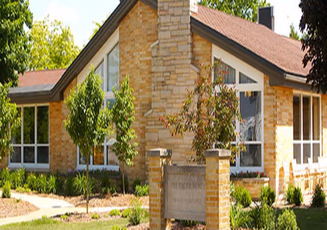 Exterior view of a single-story brick building with large windows, a stone chimney, and a landscaped garden with small trees and shrubs in front.