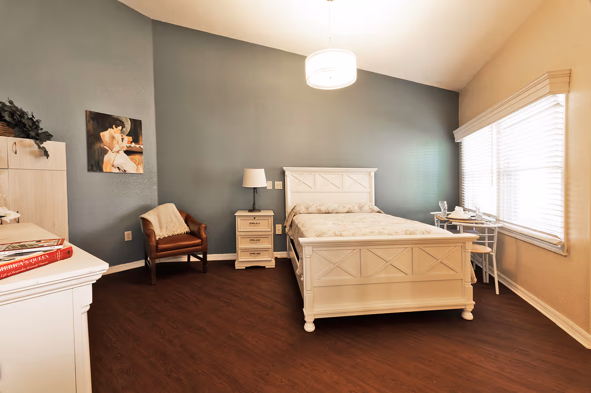 Bright furnished bedroom with a white bed, nightstand, leather armchair and dresser against a blue accent wall with a large window.