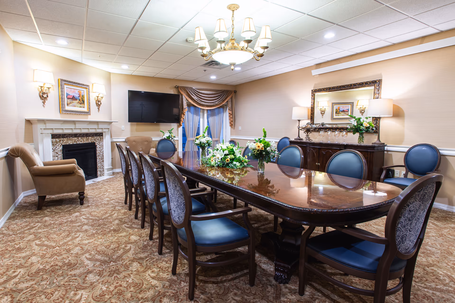 Formal dining room with a long polished table surrounded by chairs, floral centerpieces, a fireplace, wall-mounted TV, and chandelier.