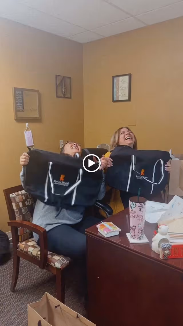Two women sitting in an office room, laughing and holding up black bags with the logo 'Revive Rehab'. The room has beige walls with framed pictures, a wooden desk with various items including a pink floral tumbler, tissue box, and a small decorative snowman. There is a patterned chair and a paper bag on the floor.