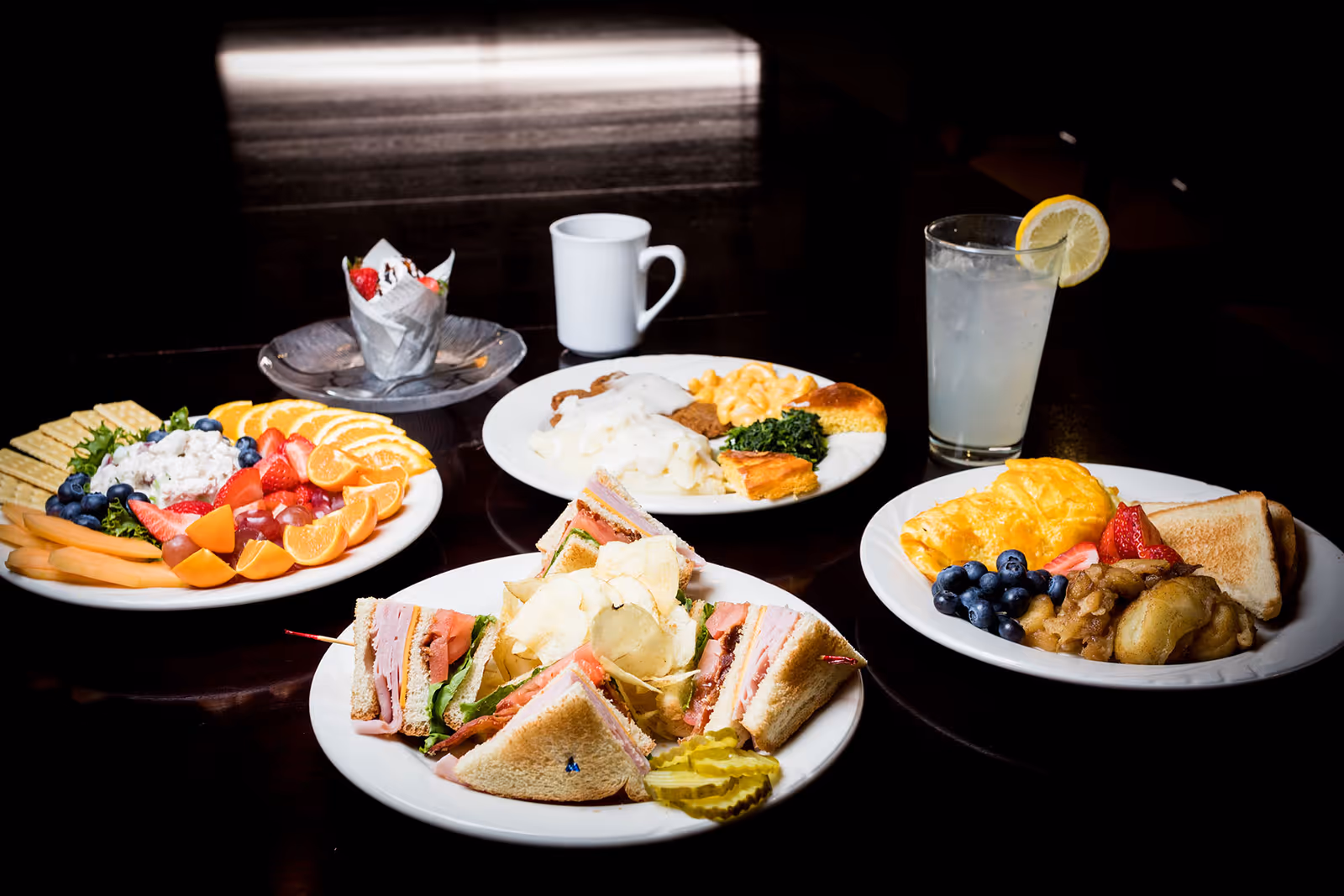 A dark wooden table set with four plates of food including a fruit and cracker platter, a club sandwich with potato chips and pickles, a breakfast plate with scrambled eggs, blueberries, strawberries, toast, and cooked apples, and another plate with mashed potatoes, gravy, macaroni and cheese, greens, and cornbread. There is also a glass of lemonade with a lemon slice and a white coffee mug on the table.