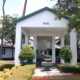 Front exterior view of a building with the number 1651 displayed above the entrance. The entrance has a covered portico supported by white columns, with a three-tiered fountain in the center of the walkway leading up to the doors. There are neatly trimmed bushes and trees on either side of the walkway.