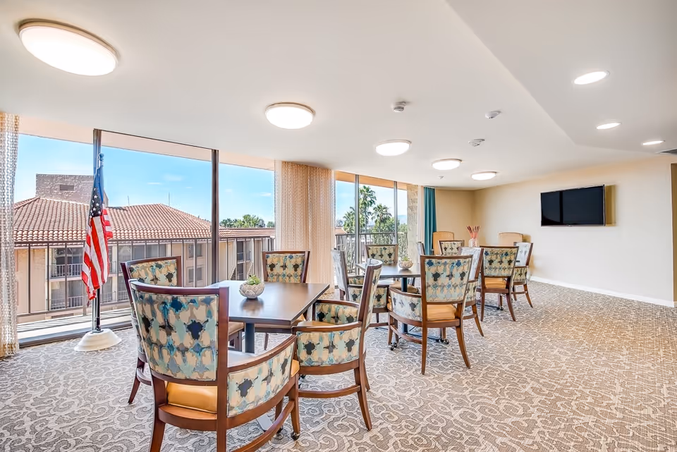 A bright and spacious common area with several wooden tables and upholstered chairs arranged neatly. Large windows let in natural light and offer a view of a building with a tiled roof and palm trees outside. An American flag stands near the windows, and a flat-screen TV is mounted on the wall. The room has patterned carpet flooring and ceiling lights.