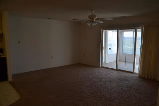 Empty living room with beige carpet, white walls, a ceiling fan with lights, and sliding glass doors leading to a sunroom or enclosed porch with windows and blinds.