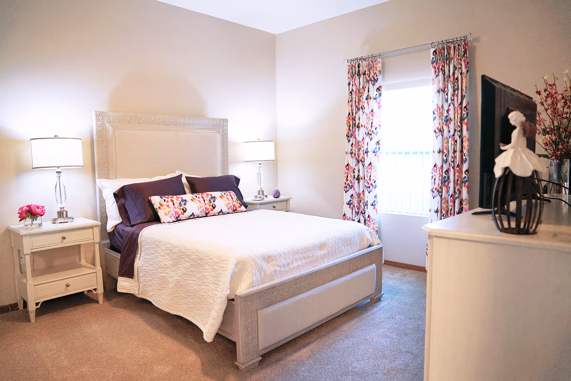 A bright and cozy bedroom with a large bed featuring a white quilt and purple pillows. There are two bedside tables with matching lamps and a small vase of pink flowers on one side. A window with floral curtains lets in natural light, and a dresser with a TV and decorative figurine is visible on the right side.