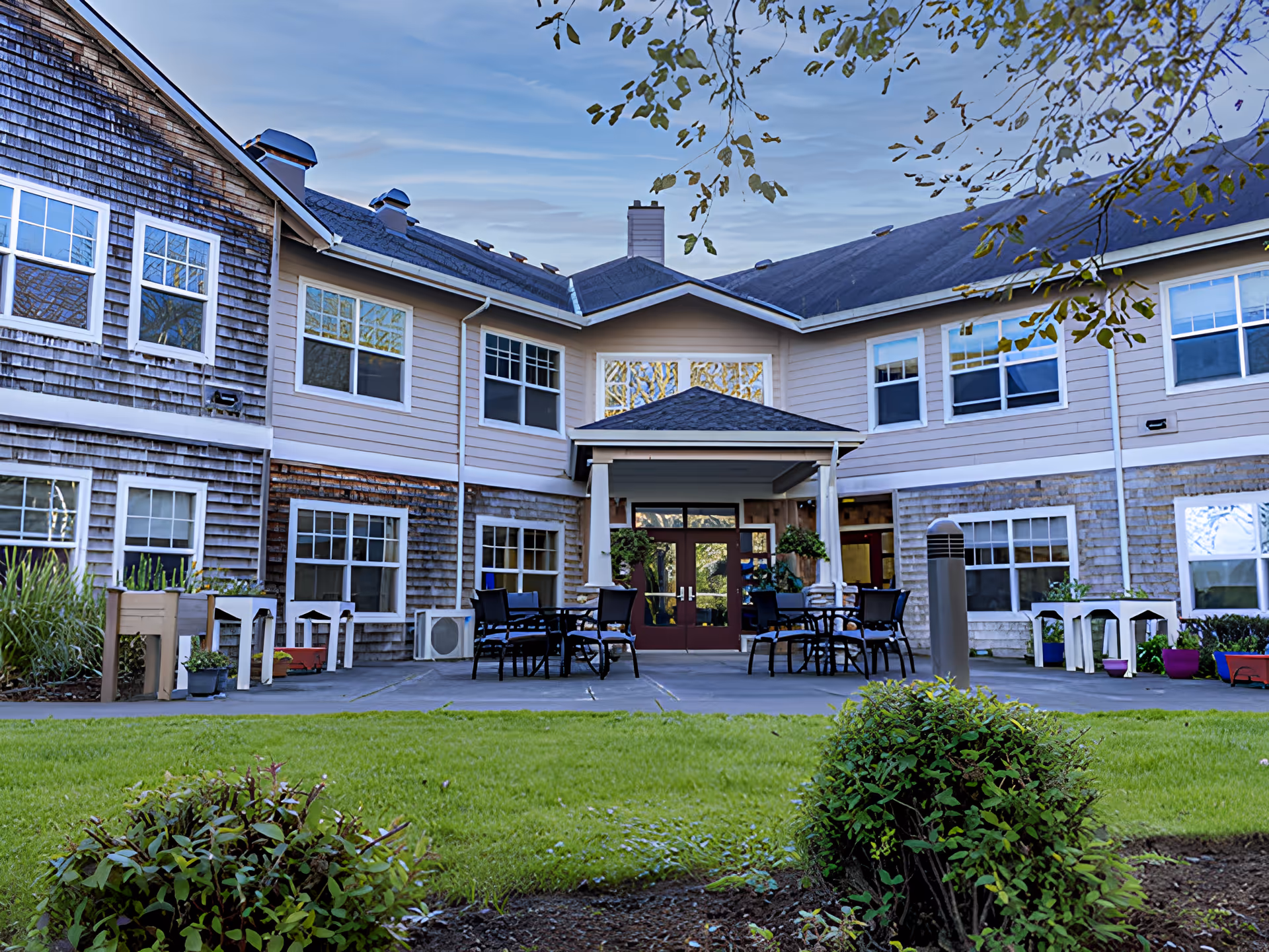Exterior view of Avamere at Seaside senior living facility showing a two-story building with multiple windows, a covered entrance with outdoor seating including tables and chairs, surrounded by green grass and shrubs under a blue sky.