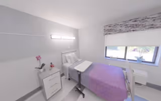 Small, bright single-occupancy room with a bed topped by a purple blanket, an overbed table, bedside cabinet with a plant, and a window.