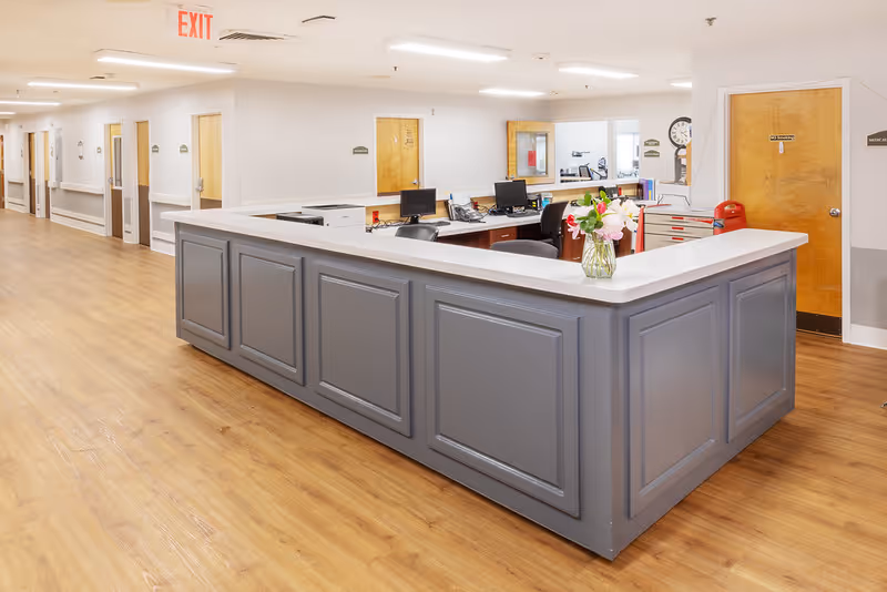 A clean and well-lit nursing station in a senior living facility with a large gray counter, several computer monitors, office chairs, and a vase of flowers on the counter. The background shows a hallway with wooden doors and an exit sign on the ceiling.