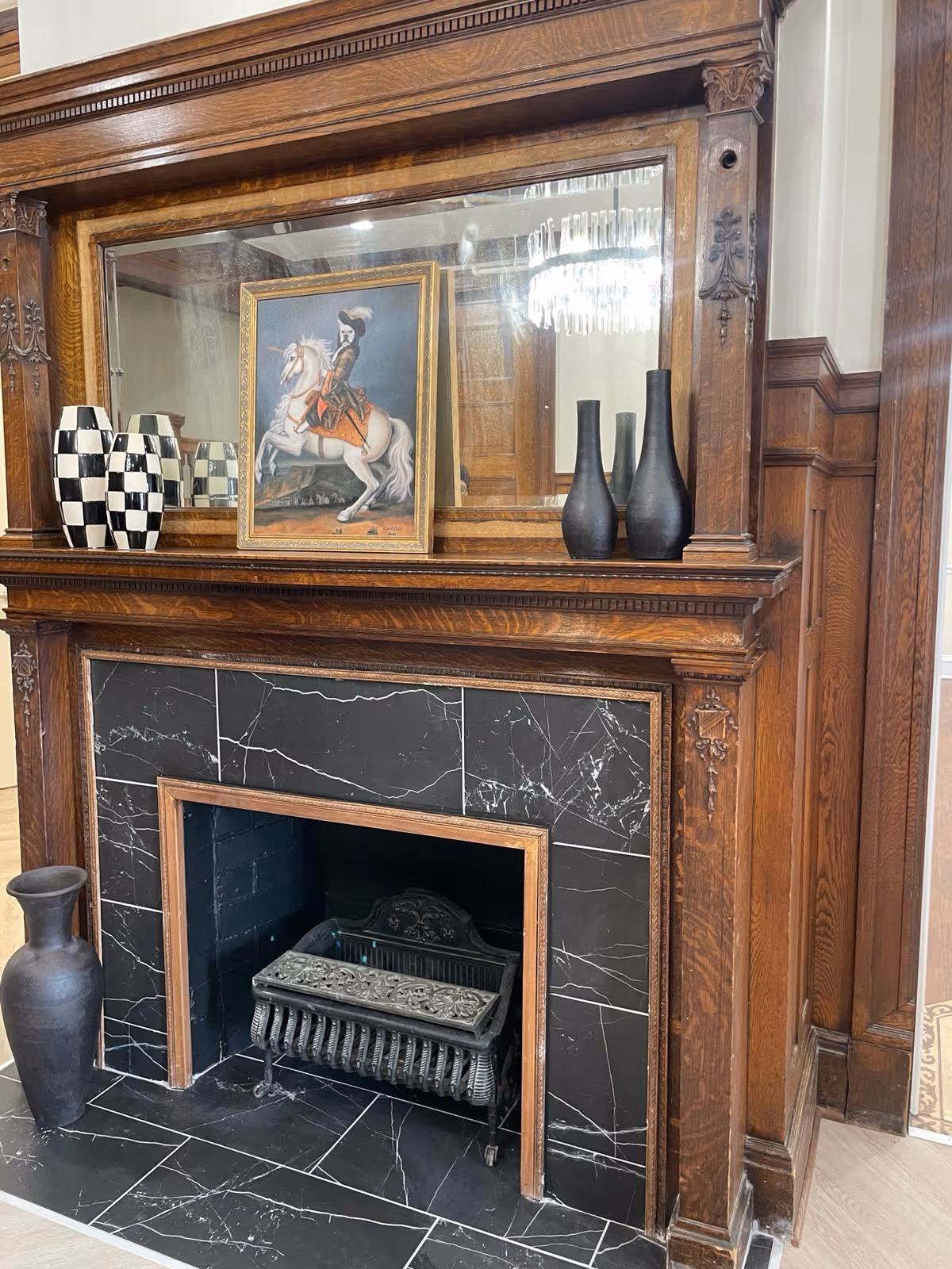 A decorative wooden fireplace with black marble tiles surrounding the firebox. On the mantel, there is a large mirror, a framed painting of a person riding a white horse, two black vases, and two black and white checkered vases. A tall black vase is placed on the floor to the left of the fireplace.